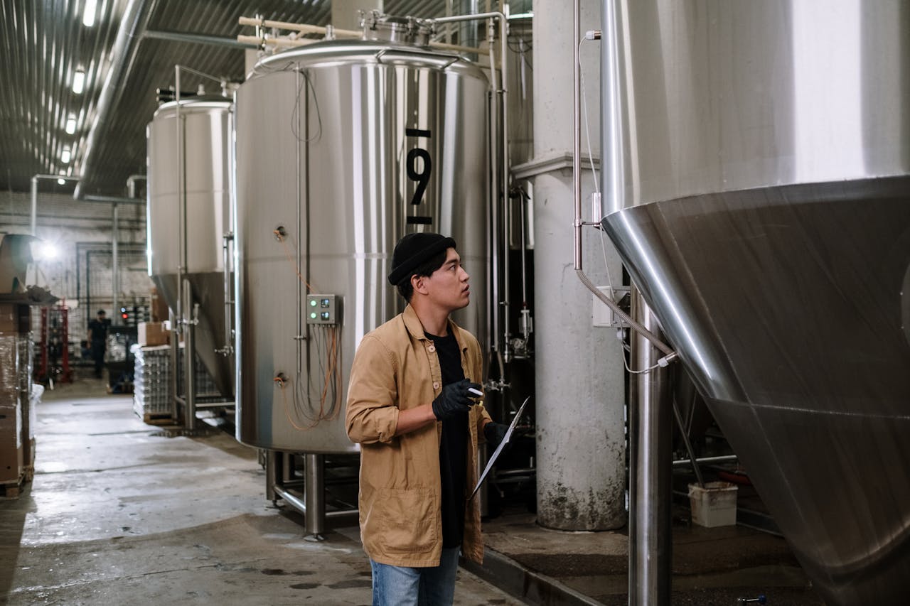Asian man inspecting stainless steel tanks in a brewery, showcasing modern industrial processes.